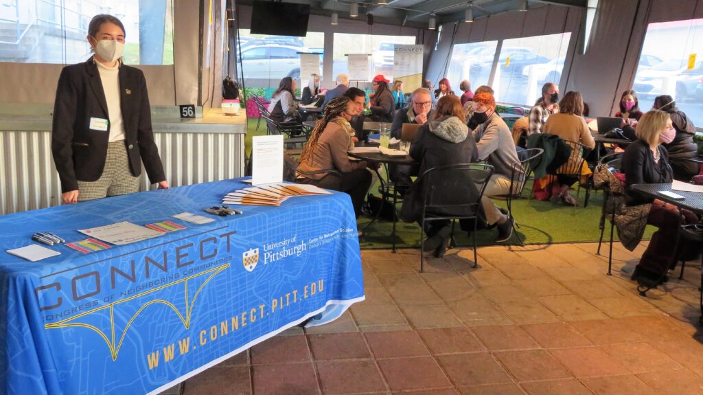 A woman stands behind a table at event with groups of people collaborating in the background