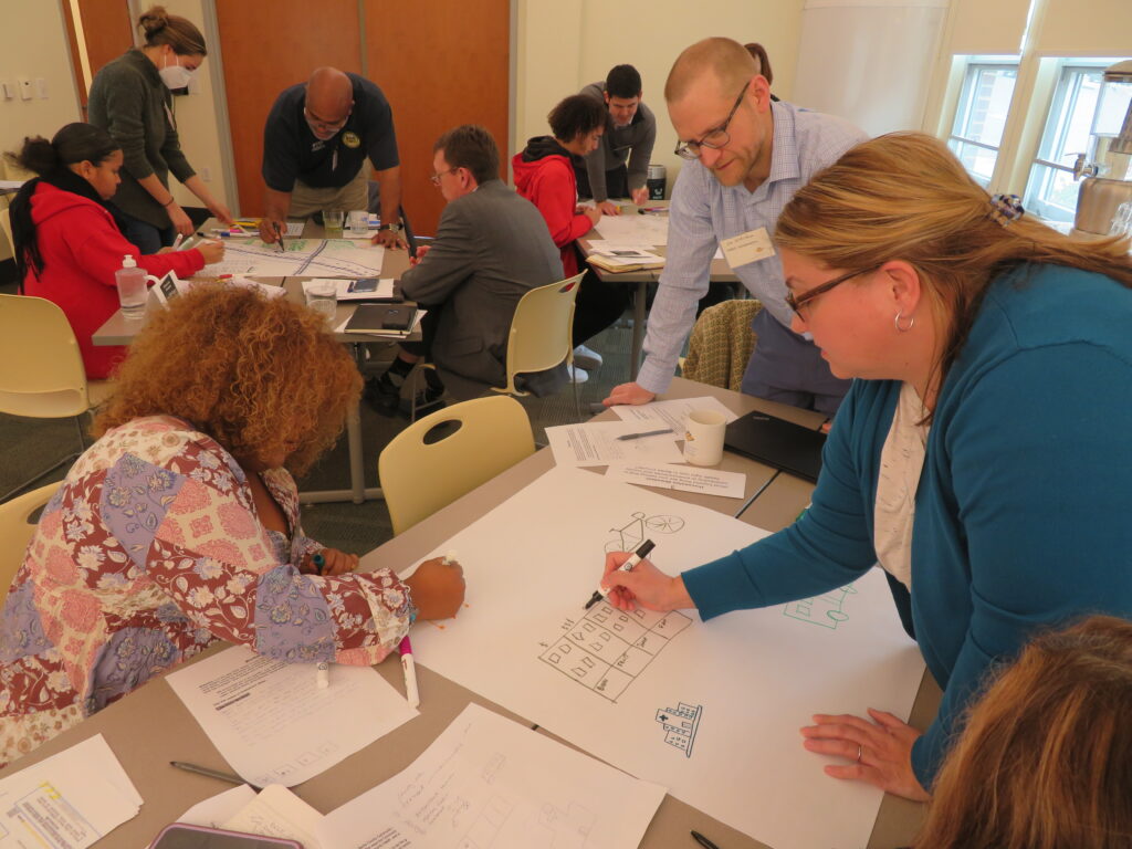 Multiple groups of leaders gather around tables to draw images of a thriving community