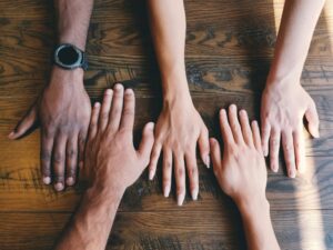 Five hands with differing skin tones lay across a table illustrating community and partnership.